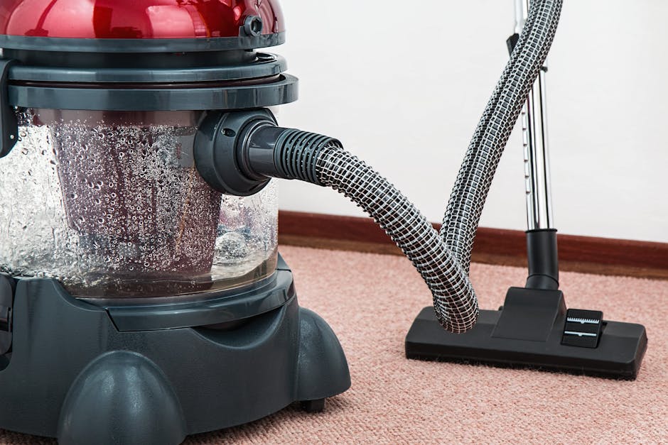 A person kneeling on a patterned carpet in a domestic setting, opening the lid of a yellow portable vacuum cleaner with a black hose, preparing for surface cleaning. The individual is dressed in casua