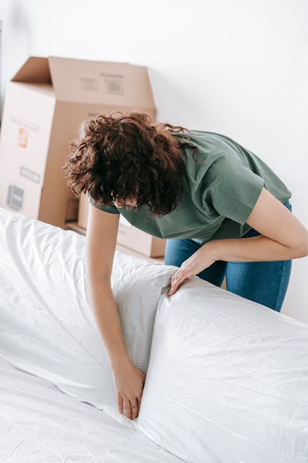 A woman with curly brown hair wearing a green t-shirt is performing surface cleaning on a large white sofa in a well-lit room. She is carefully smoothening and wiping the fabric of the sofa, which app