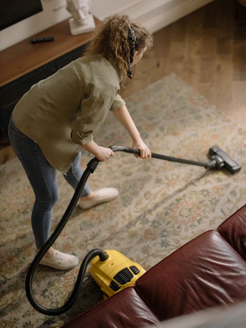 A woman with curly hair, dressed in a light green jacket, dark jeans, and white shoes, is using a vacuum cleaner on a patterned area rug in a living room. The vacuum is yellow with a black hose, and s